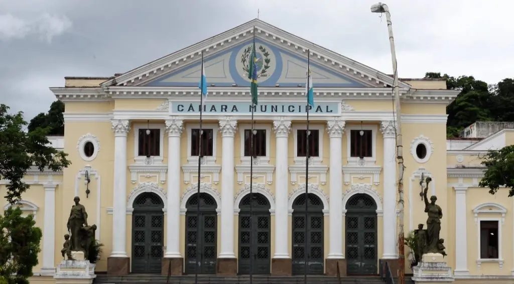 Vista frontal de edifício neoclássico com frontão triangular, denominado “Câmara Municipal”. A fachada apresenta seis janelas em arco com detalhes intrincados, três mastros com bandeiras e estátuas nos cantos da entrada. O céu está nublado.