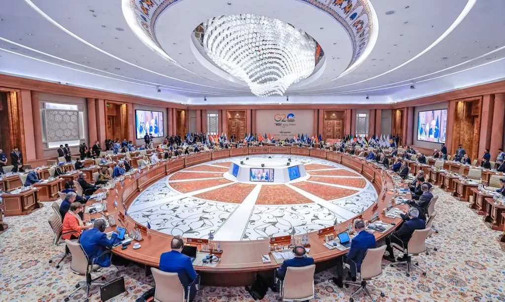 Encontro do G20 em 2023 na Índia. Na imagem, reunião oficial da cúpula, com representantes de diversos países sentados em volta de uma mesa redonda gigante, aberta no meio.