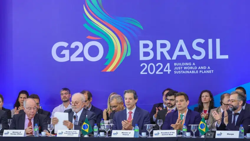 Na imagem, o atual presidente Lula e ministros do seu governo sentados em uma mesa de pronunciamento.