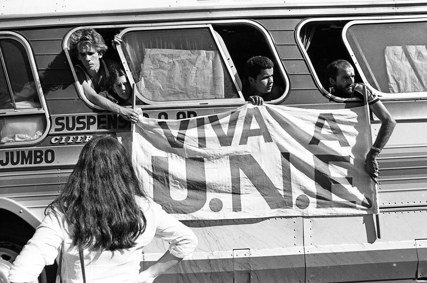 a imagem mostra pessoas dentro de um ônibus segurando uma bandeira "VIVA A UNE"