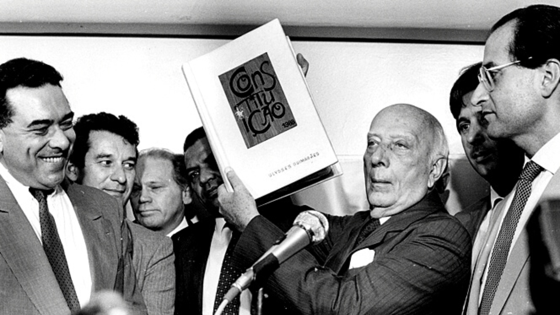 Ulysses Guimarães segurando a Constituição Federal de 1988, apresentando documento que garante a cobrança progressiva do Imposto sobre a Renda.