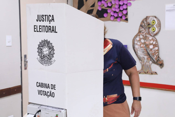 A imagem apresenta uma cabine de votação pelo lado de fora. é possível ver o nome "Justiça Eleitoral" e o símbolo da República. Uma pessoa que está dentro da cabine, vota. não é possível ver seu rosto.