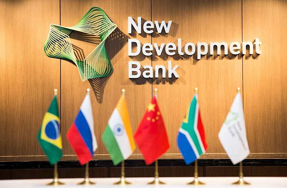 A imagem mostra pequenas bandeiras dos países que forma o BRICS lado a lado. Brasil, Rússia, índia, China e África do Sul, ao lado delas a bandeira do New Development Bank. Ao fundo o nome e o símbolo do New Development Bank. 