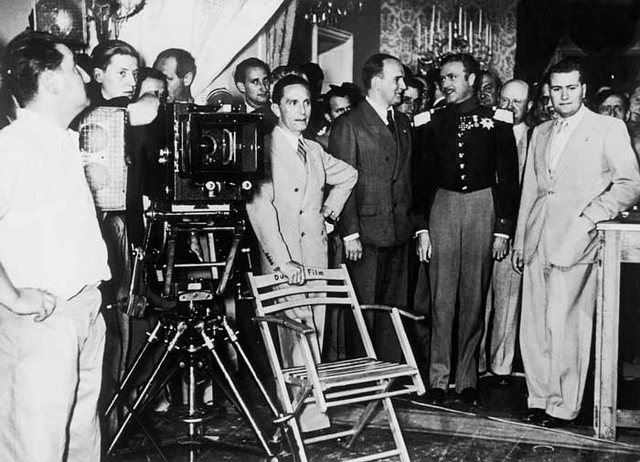 Homens vestidos formalmente reunidos em um set de filmagem e uma câmera antiga em ação.