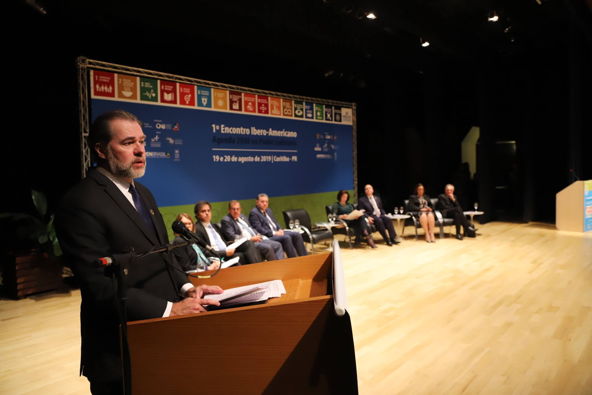 Na imagem, pessoas reunidas em frente a painel com os dizeres 1º encontro Ibero-americano. Conteúdo sobre Agenda 2030.