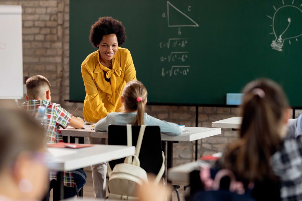 Foto de uma sala de aula com um quadro ao fundo e a professora atenta às demandas dos alunos para promover uma educação de qualidade