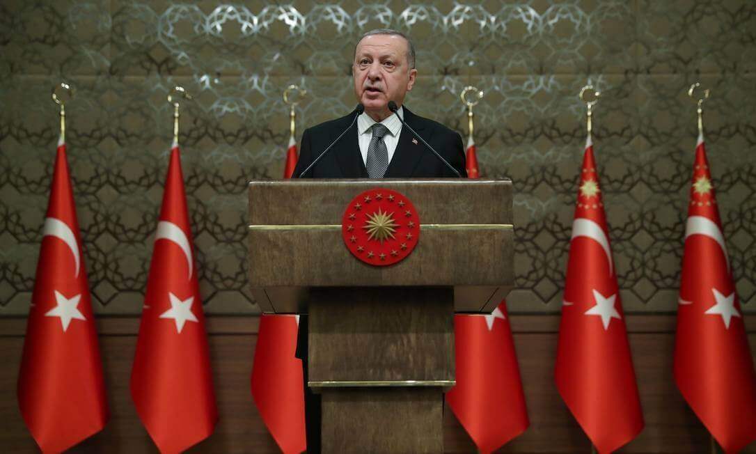 Na imagem, presidente da Turquia em pé, durante discurso, rodeado de bandeiras do país.