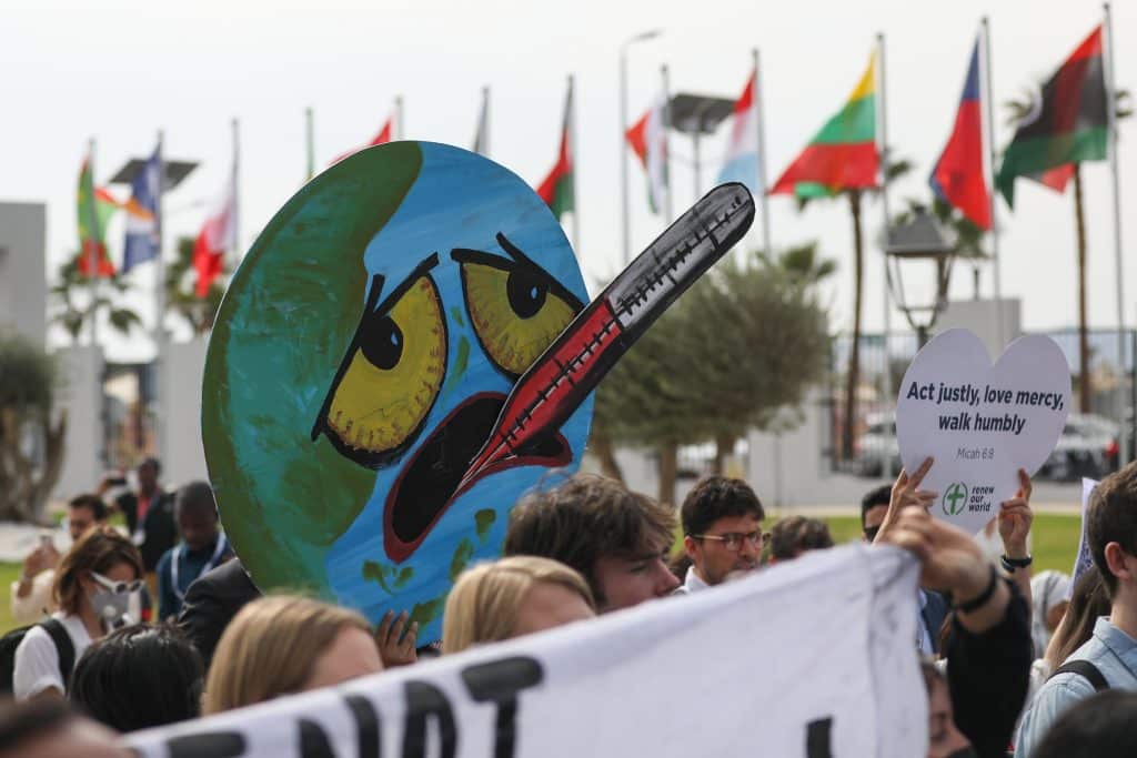 A imagem é uma foto que mostra uma manifestação. As pessoas seguram cartazes, em destaque, o desenho do planeta Terra com um termômetro na boca. O desenho tem olhos cansados e o termômetro indica febre.