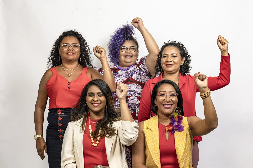 Alt Tag: Foto com 5 mulheres de diversas etnias, que estão com a mão esquerda levantada simbolizando a luta pela igualdade de gênero. A Lei das Eleições é parte importante na promoção do acesso aos espaço políticos.