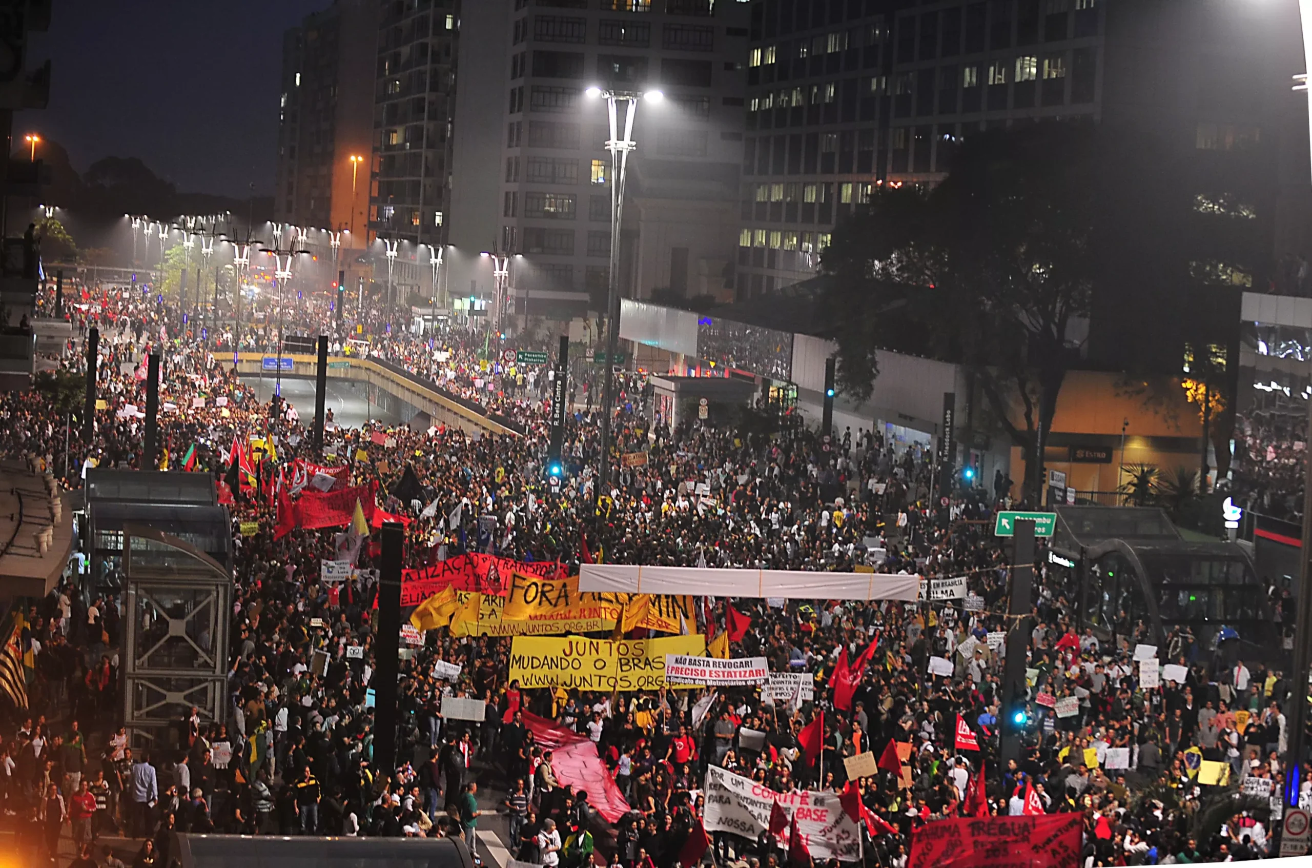 A imagem apresenta a Avenida Paulista lotada de manifestantes. Eles seguram placas e faixas. Uma delas, tem escrita a seguinte frase " Juntos mudando o Brasil". 