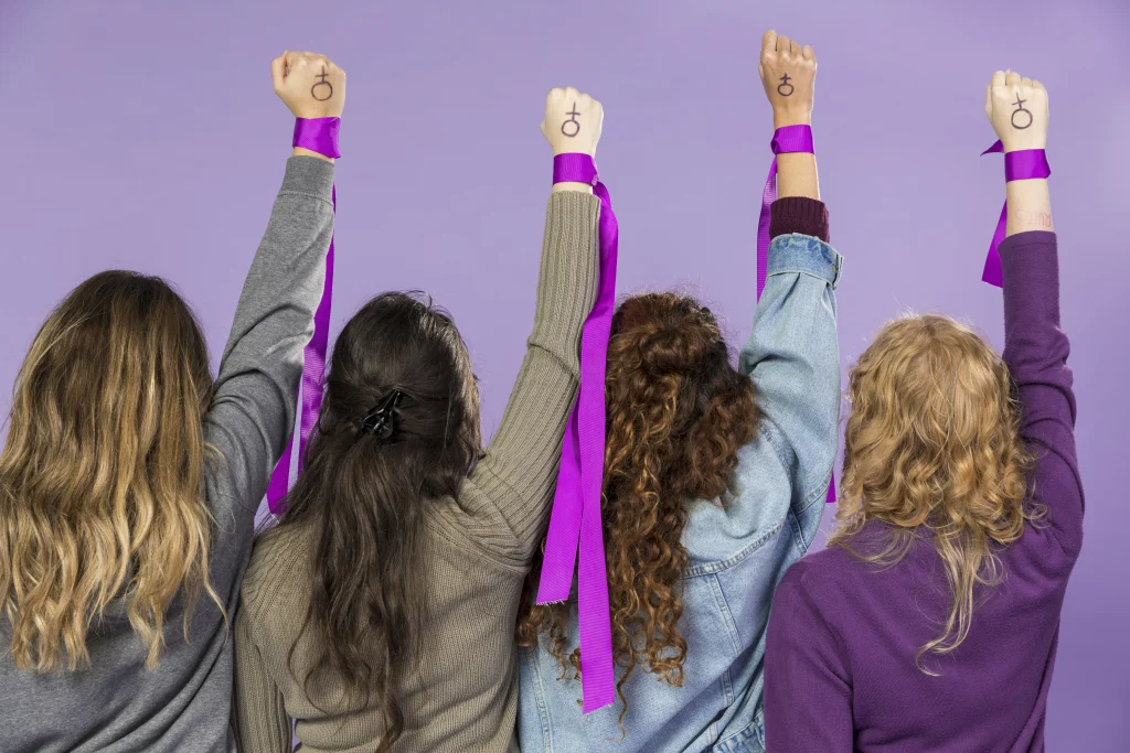  Imagem com quatro mulheres com a mão esquerda levantada com o símbolo das mulheres para simbolizar o combate à desigualdade de gênero 