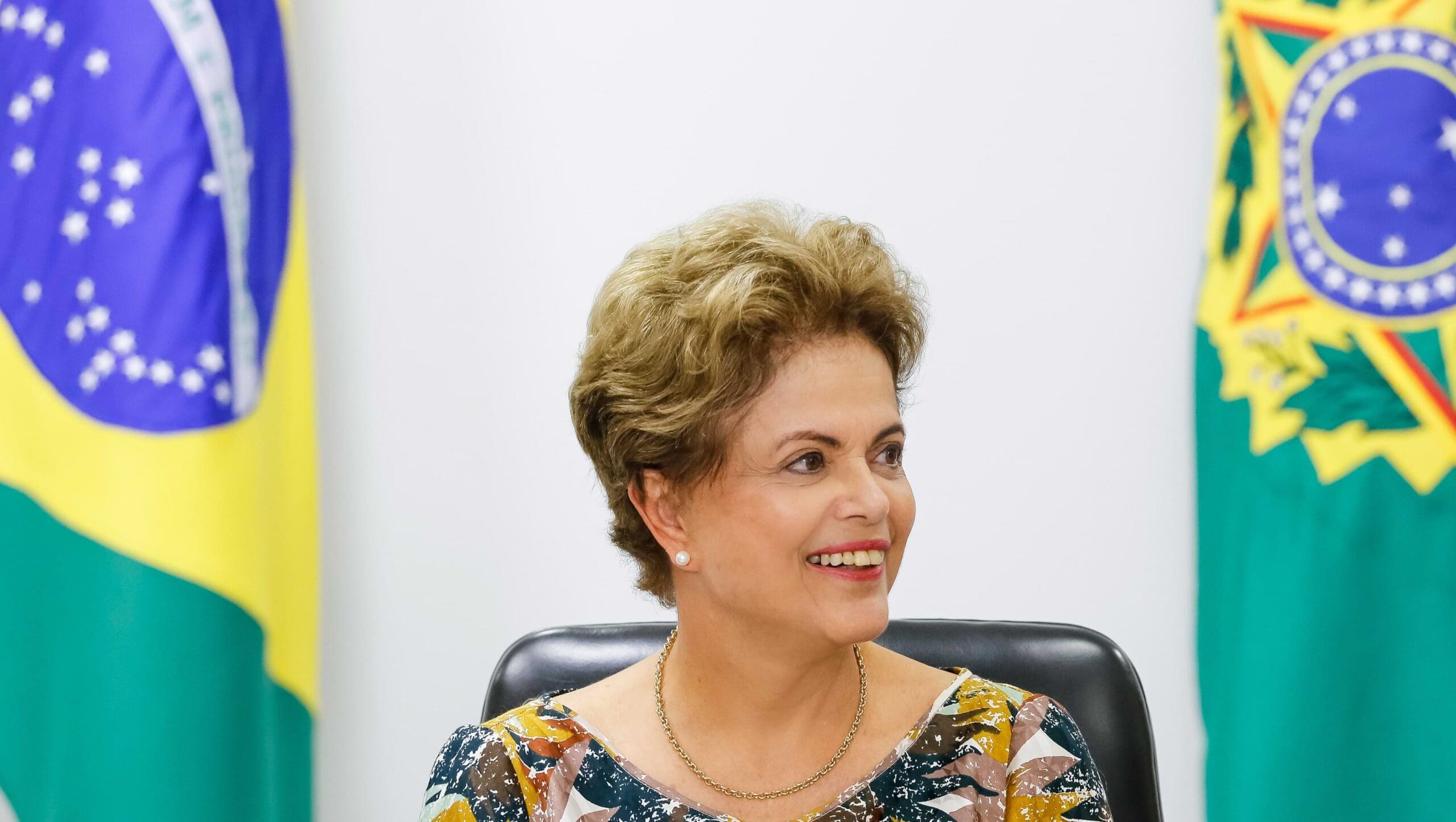 Dilma sentadana frente da bandeira do Brasil.