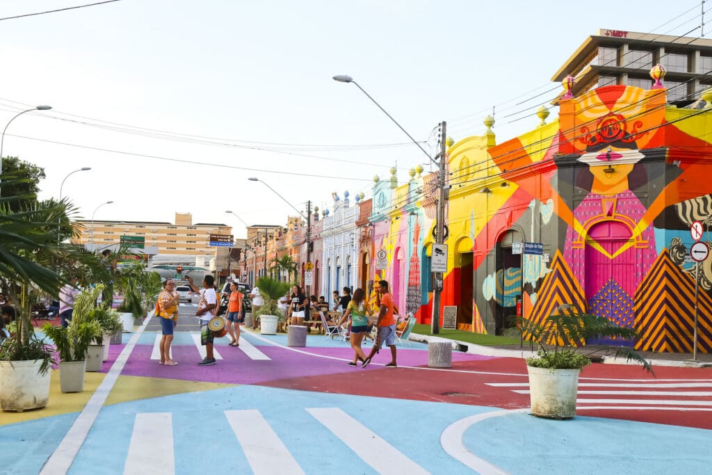 A imagem apresenta um rua. Ela tem construções pequenas no lado direito e plantas do lado esquerdo. A construções são coloridas e o chão também é pintado 