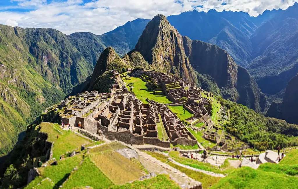 A imagem mostra a cidade de Machu Picchu, localizada no alto de uma montanha. Algumas estruturas ainda estão intactas. Ao fundo é possível ver uma cadeia de montanhas.