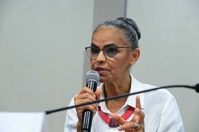 Marina Silva está centralizada na imagem, com cabelo preso, vestindo blusa branca e usando um colar vermelho. Ela está falando, com um microfone na mão.