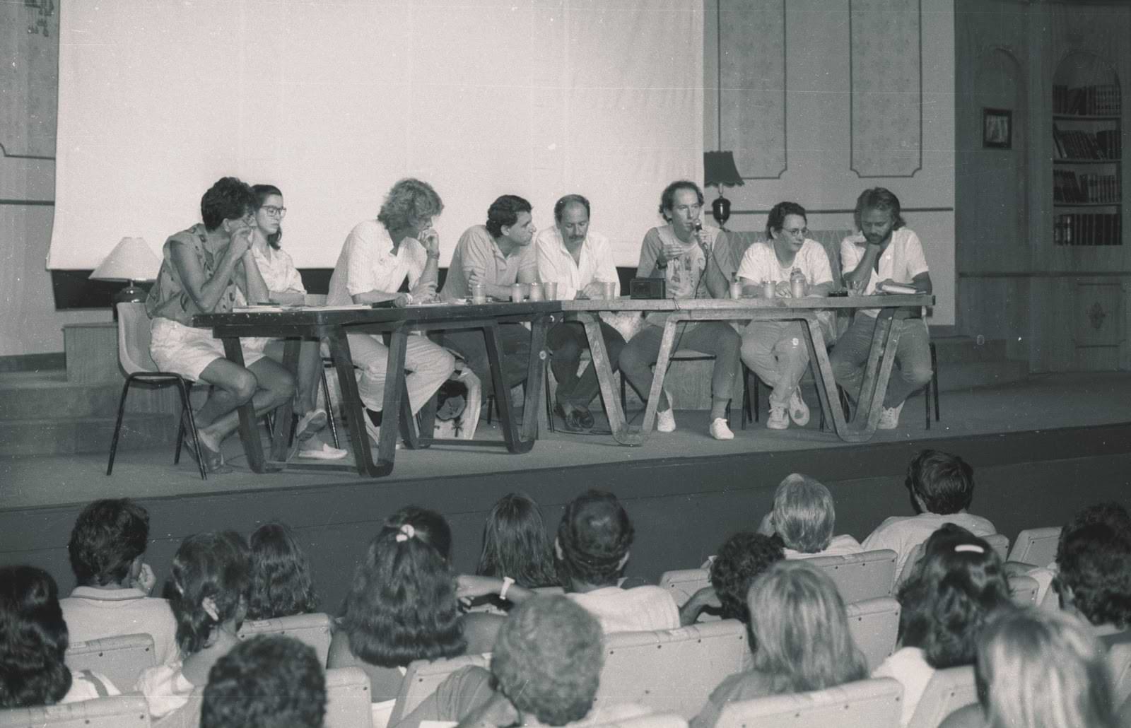 Imagem da decada de 80 de uma assembleia com um palco, no palco atrás de uma mesa encontram-se 5 homens e 3 mulheres.