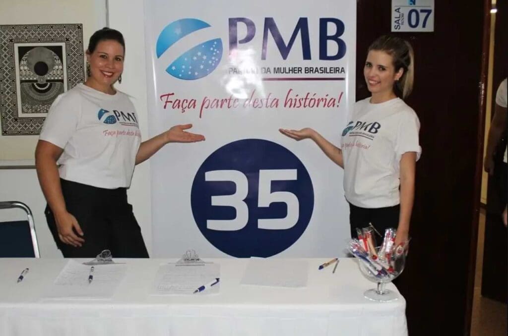 Descrição da imagem: Duas mulheres vestidas com a camisa branca do partido, apontando para o cartaz onde tem escrito “PMB - Partido da Mulher Brasileira. Faça parte desta história!”. 