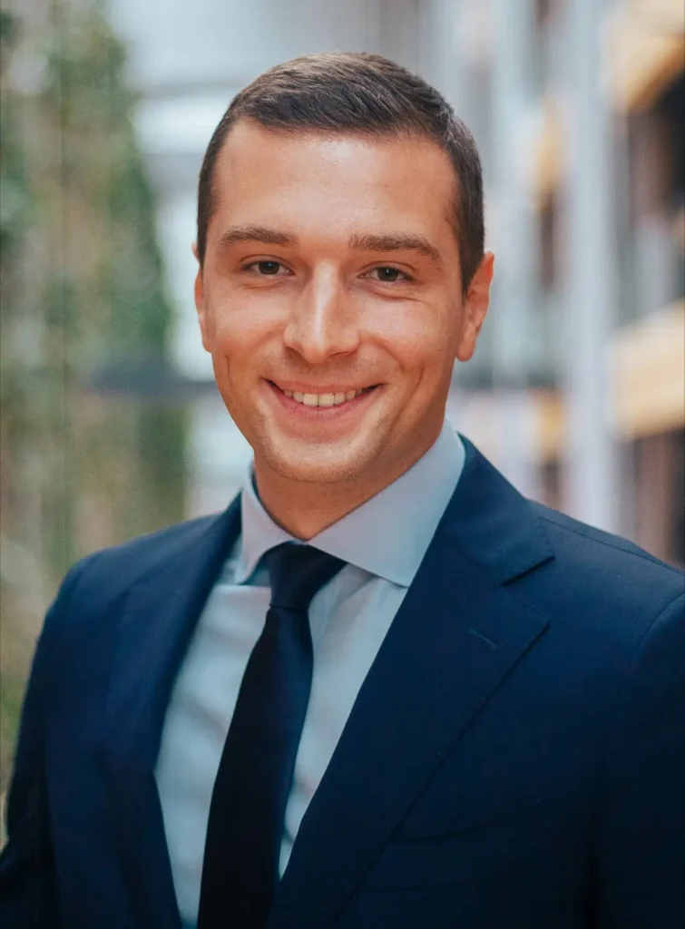 homem branco jovem sorridente, com cabelos curtos e utilizando um terno azul