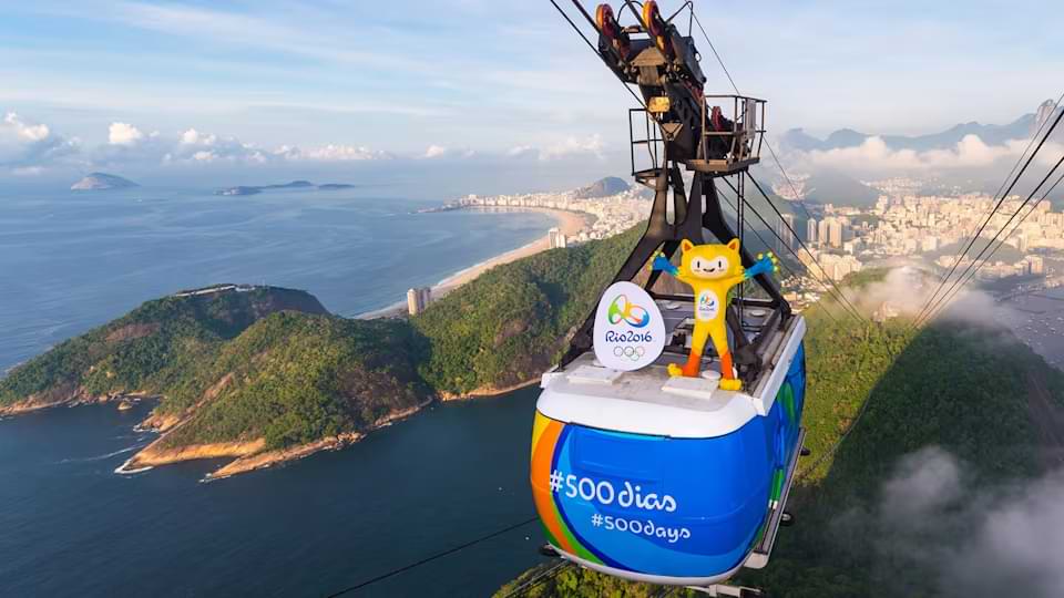 Alt text: Mascote das Olimpíadas semelhante a um gato, representando a onça pintada, em cima do Bondinho do Pão de Açúcar, no fundo a Praia da Urca e a cidade do Rio de Janeiro.