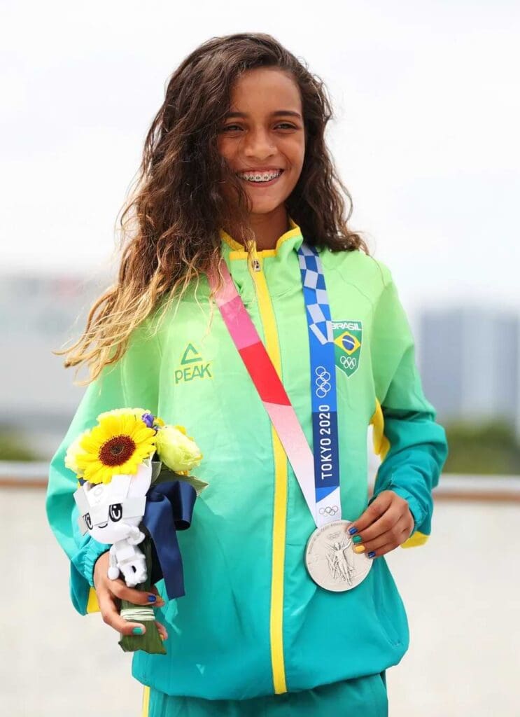 Menina negra com cabelos ondulados grandes, utilizando casaco com as cores verde e amarelo, segurando um buquê e carregando uma medalha de prata no pescoço.