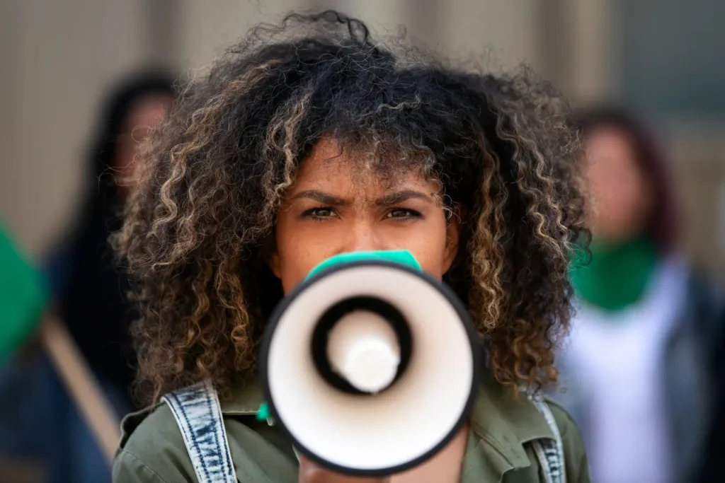 Uma pessoa com cabelos cacheados segura um megafone e olha diretamente para a câmera durante um protesto ao ar livre. Eles estão usando uma bandana verde no pescoço e uma camisa verde. O fundo mostra outras pessoas, um pouco desfocadas, também participando do protesto. Perfeito para fotos de banco de imagens.