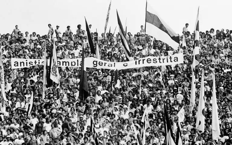 Imagem em preto e branco de torcedores com diversas bandeiras do time Corinthians. No meio da torcida é estendida uma faixa com os dizeres: "anistia ampla geral e irrestrita".