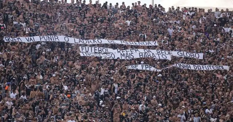 Imagem colorida de torcedores que vestem camisetas em preto e branco do time Corinthias e estendem quatro faixas brancas com escrito em preto. De baixo para cima, a primeira faixa diz: "Quem vai punir o ladrão da merenda?"; a segunda faixa diz: "Futebol refém da Globo"; a terceira diz: "Ingresso mais barato" e a quarta diz: "CBF FPF vergonha do futebol".