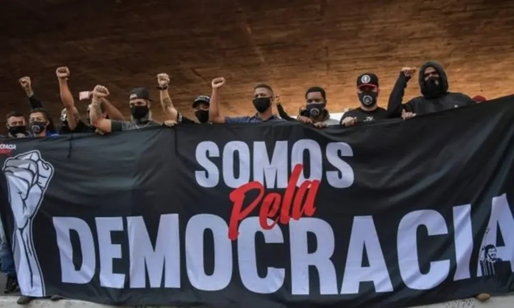 Imagem colorida contendo dez homens jovens, que vestem preto e levantam as mãos com o punho fechado, todos usam uma máscara preta e três usam bonés pretos. Eles estendem uma faixa com os dizeres: "Somos pela democracia". A faixa é preta e o escrito está em branco, sendo somente a palavra "pela" destaca em vermelho bem ao centro da faixa. Na faixa, no canto esquerdo da imagem, está desenhado, em branco, uma mão com o punho fechado.