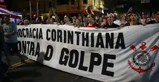 Imagem colorida contendo pessoas em uma manifestação segurando uma faixa branca com os dizeres em preto: "Democracia corinthiana contra o golpe". Na faixa, do lado direito da imagem contém o símbolo do clube Corinthians.