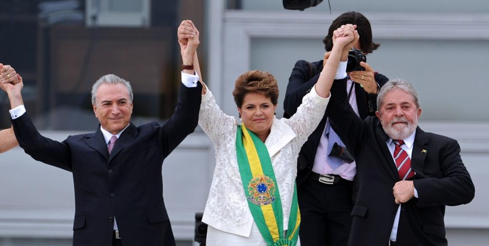 A imagem mostra uma foto de Michel Temer, Dilma Rousseff e Luiz Inácio Lula da Silva. Os três estão lado a lado, de mãos dadas e elevadas acima da cabeça.