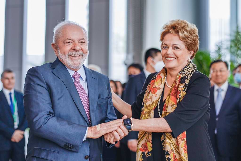 Foto de Lula, homem branco baixo idoso, com cabelos curtos e arba curta branca usando um terno, cumprimentando Dilma, mulher de mesma estatura de Lula, cabelos curtos loiros, usando um vestido preto.