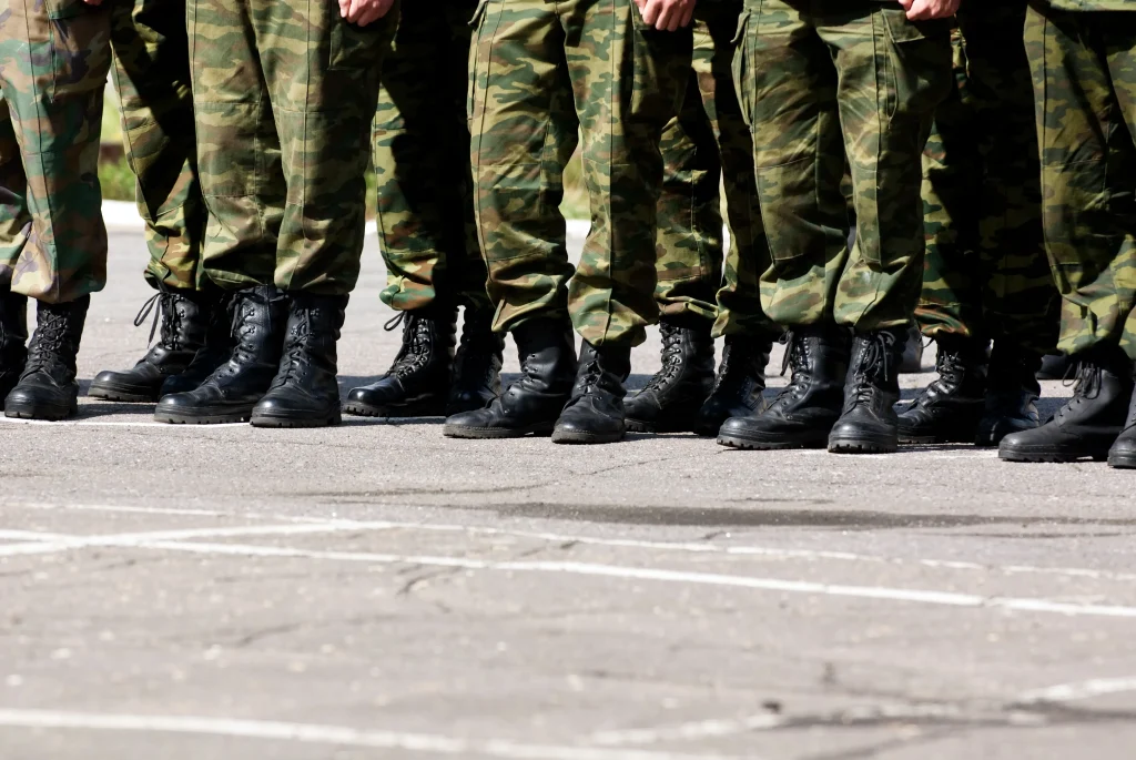 A imagem mostra a parte inferior das pernas de várias pessoas vestidas com uniformes militares camuflados e botas pretas, alinhadas em formação sobre uma superfície pavimentada. Texto sobre neofascismo.