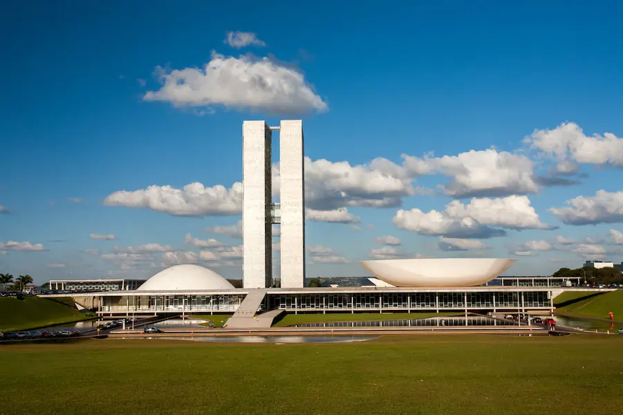 A imagem mostra a fachada do Congresso Nacional do Brasil, localizado em Brasília. O edifício icônico apresenta duas torres altas e simétricas no centro, ladeadas por duas cúpulas distintas: a cúpula menor e arredondada à esquerda, que abriga o Senado Federal, e a cúpula maior e invertida à direita, que abriga a Câmara dos Deputados. O céu azul com algumas nuvens brancas complementa a grandiosidade da arquitetura modernista do prédio, projetado por Oscar Niemeyer. Texto sobre vagas no Congresso.