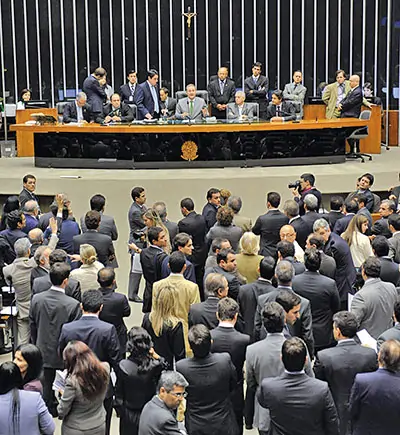 A imagem mostra uma sessão parlamentar em um plenário lotado. Diversos políticos estão reunidos, alguns de pé e outros sentados, enquanto uma pessoa está presidindo a sessão ao fundo, na mesa principal. A cena retrata uma discussão ou votação, com um grande número de pessoas envolvidas, possivelmente em uma câmara legislativa ou congresso. O ambiente é formal, com trajes sociais predominando entre os presentes. Texto sobre as vagas no Congresso.