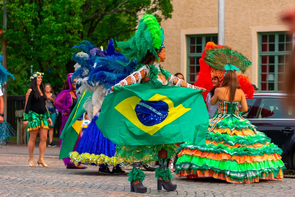 A imagem mostra um grupo de pessoas participando de uma festa ou desfile com temática brasileira. No centro, uma mulher usa uma fantasia colorida, adornada com penas verdes e azuis, e segura uma grande bandeira do Brasil. Outras pessoas ao redor também estão vestidas com trajes vibrantes e elaborados, típicos de festas como o Carnaval. O ambiente ao ar livre e as expressões festivas indicam uma celebração cultural, com muita cor e alegria. TExto sobre identidade nacional