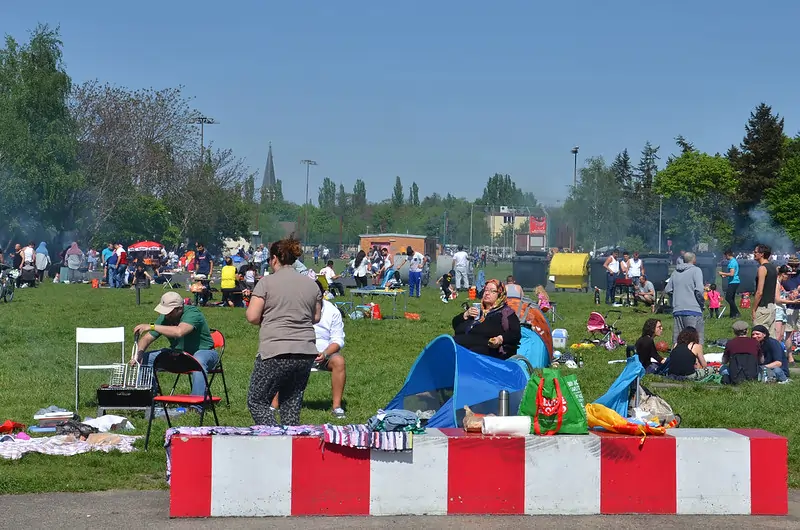 Parque cheio de pessoas aproveitando um dia ensolarado. Várias famílias e grupos de amigos estão fazendo piqueniques e churrascos ao ar livre. Algumas pessoas estão sentadas em cadeiras e no chão, enquanto outras estão conversando em pé. No primeiro plano, há uma mesa com vários itens e alimentos. Ao fundo, árvores verdes e um céu claro completam a paisagem.