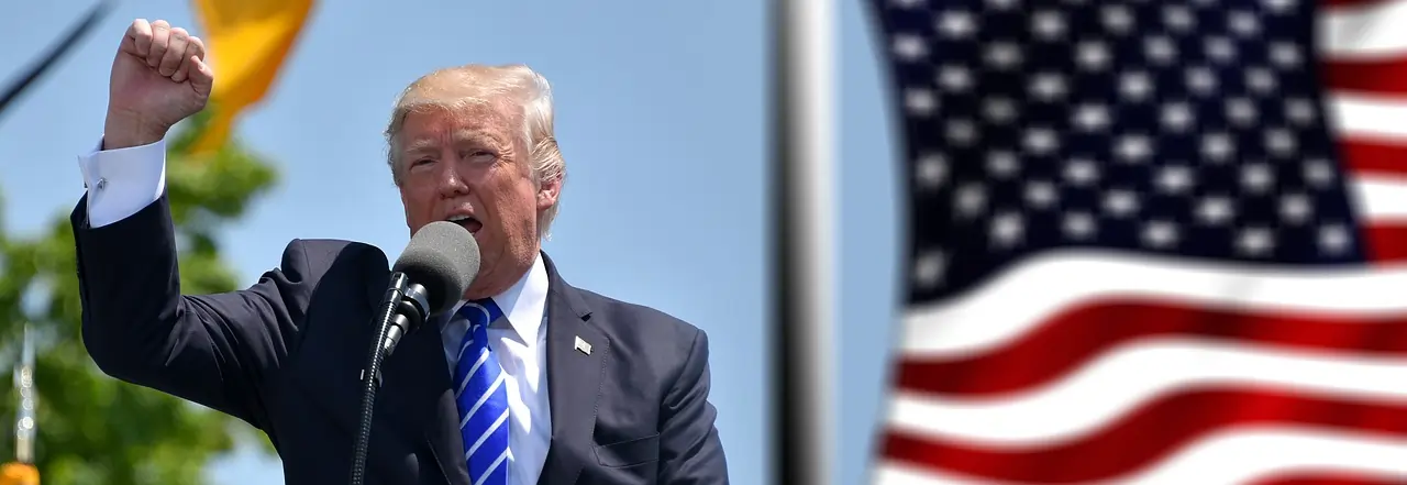 Foto de Donald Trump discurssando em um palanque com a mão erguida com a bandeira dos EUA atrás.