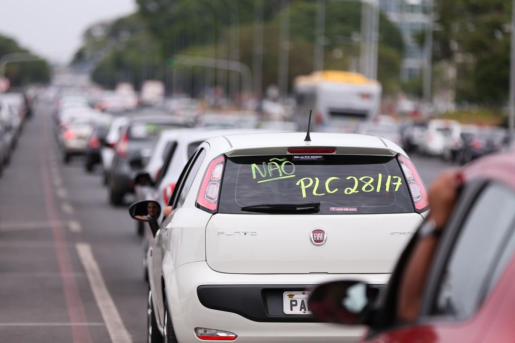 Carros enfileirados em protesto. No vidro de um carro está escrito "Não PLC 28/17".