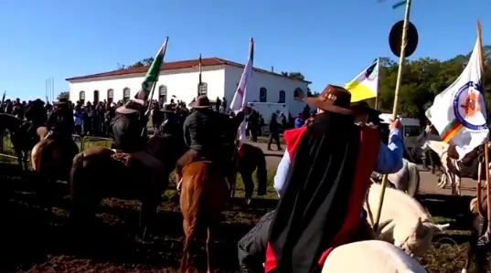 Diversos homens estão montados a cavalo e de costas para quem vê, e de frente para um casarão branco. Texto sobre a Semana Farroupilha.