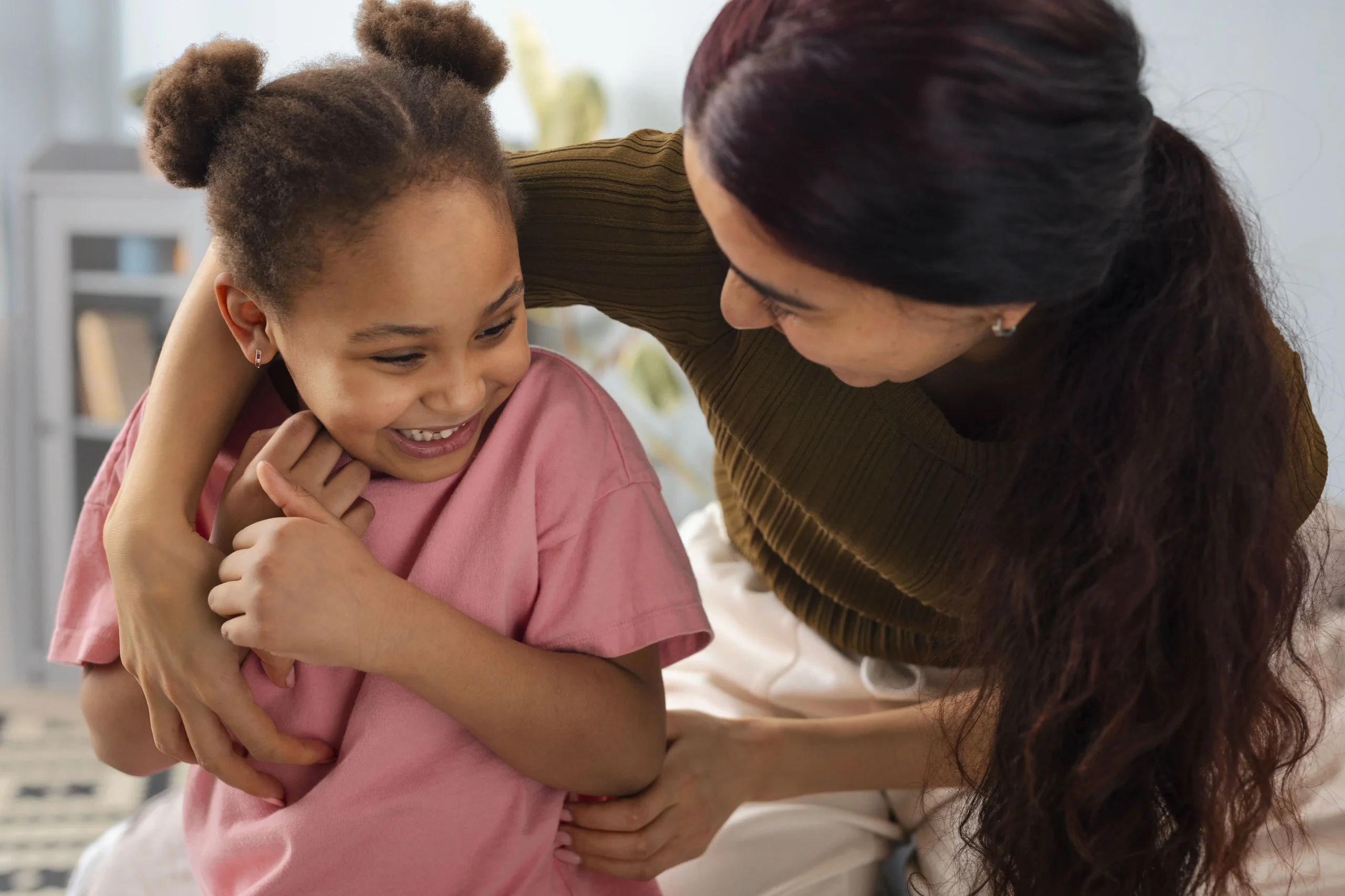 Um adulto e uma criança estão sorrindo ao brincar. A criança veste uma blusa rosa e o adulto uma blusa branca. Estão em um ambiente interno, possivelmente uma casa.