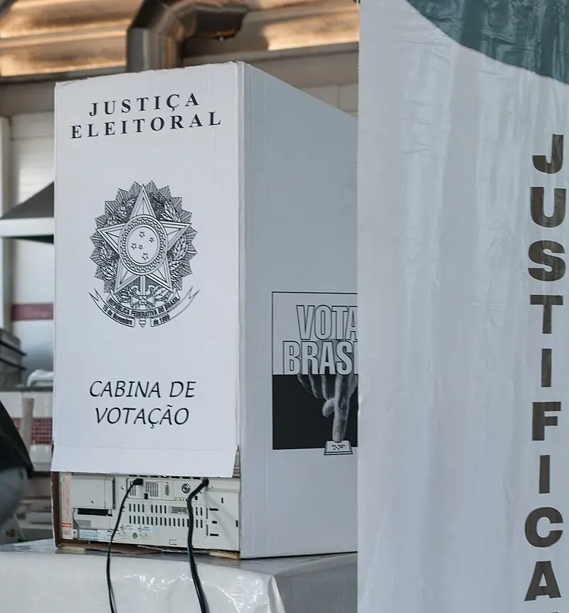 Fotografia mostrando a cabine de votação em uma seção eleitoral, com brasão da Justiça Eleitoral. Texto sobre horário de votação.