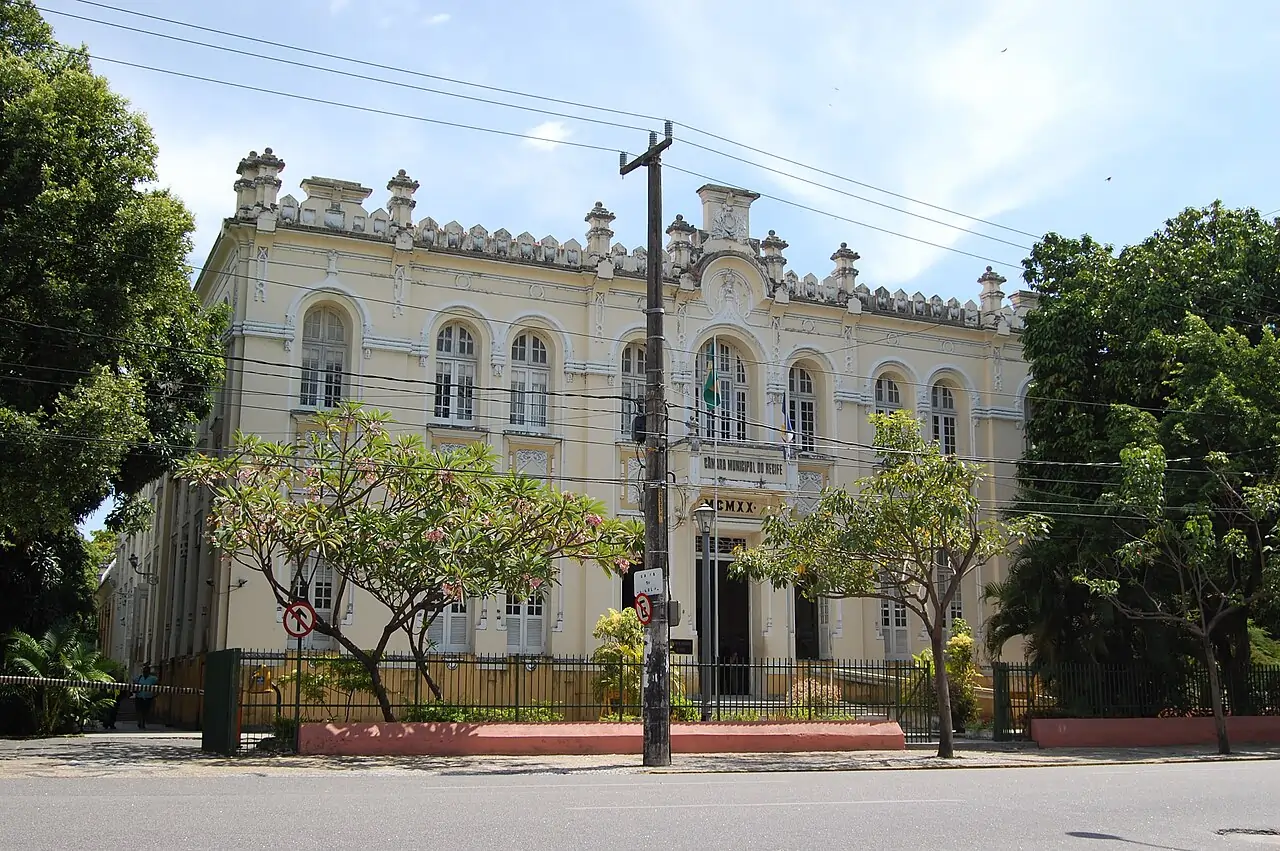 Fachada de um prédio com arquitetura antiga, na cor bege, com dois andares, atrás de cabos de energia elétrica e com árvores à direita, esquerda, e à frente, sob céu azul]. Texto sobre quantos vereadores são eleitos por cidade.