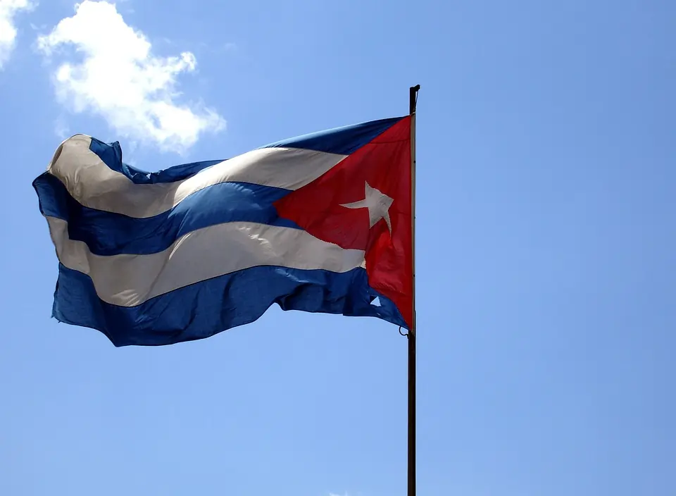 A imagem mostra a bandeira de Cuba tremulando contra um céu azul claro e  com poucas nuvens. A bandeira, composta por cinco listras horizontais alternadas em azul e branco, possui um triângulo vermelho na lateral esquerda, que contém uma estrela branca de cinco pontas no centro. Texto: A crise dos mísseis de Cuba: os 13 dias à beira da guerra nuclear