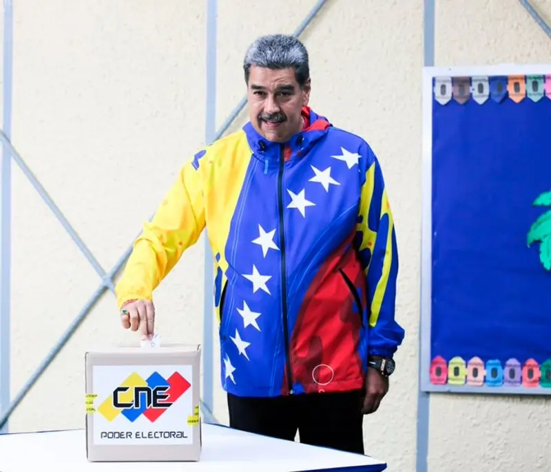 Nicolas Maduro, com um casaco com a imagem da bandeira venezuelana. Possui idade um pouco avançada, cabelo curto grisalho mesclado com preto, bigode espesso, de cor parda clara. Na foto, tem um meio sorriso. Texto: Entenda o conceito de autocracia eleitoral