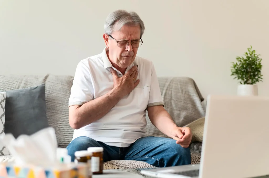 Um homem branco, de óculos, olhos fechados, cabelos brancos, com a mão no peito em sinal de dor. Está sentado e um sofá, e parece ter um computador em uma mesa baixa à sua frente. Doenças cardiovasculares, que causam dor no peito, são doenças crônicas não transmissíveis.