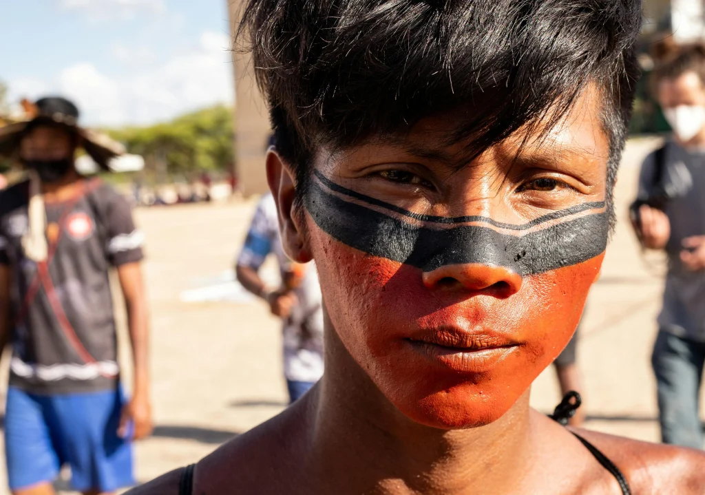 Rapaz indígena com rosto pintados em linhas pretas e uma porção do rosto, abaixo do nariz, em vermelho. Texto: APIB: entenda o que são e o que defendem