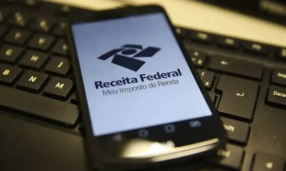 Imagem de um celular sob um teclado de computador com a imagem da Receita Federal (Meu Imposto de Renda) na tela. Texto: Tributação das apostas on-line no Brasil: como funciona?